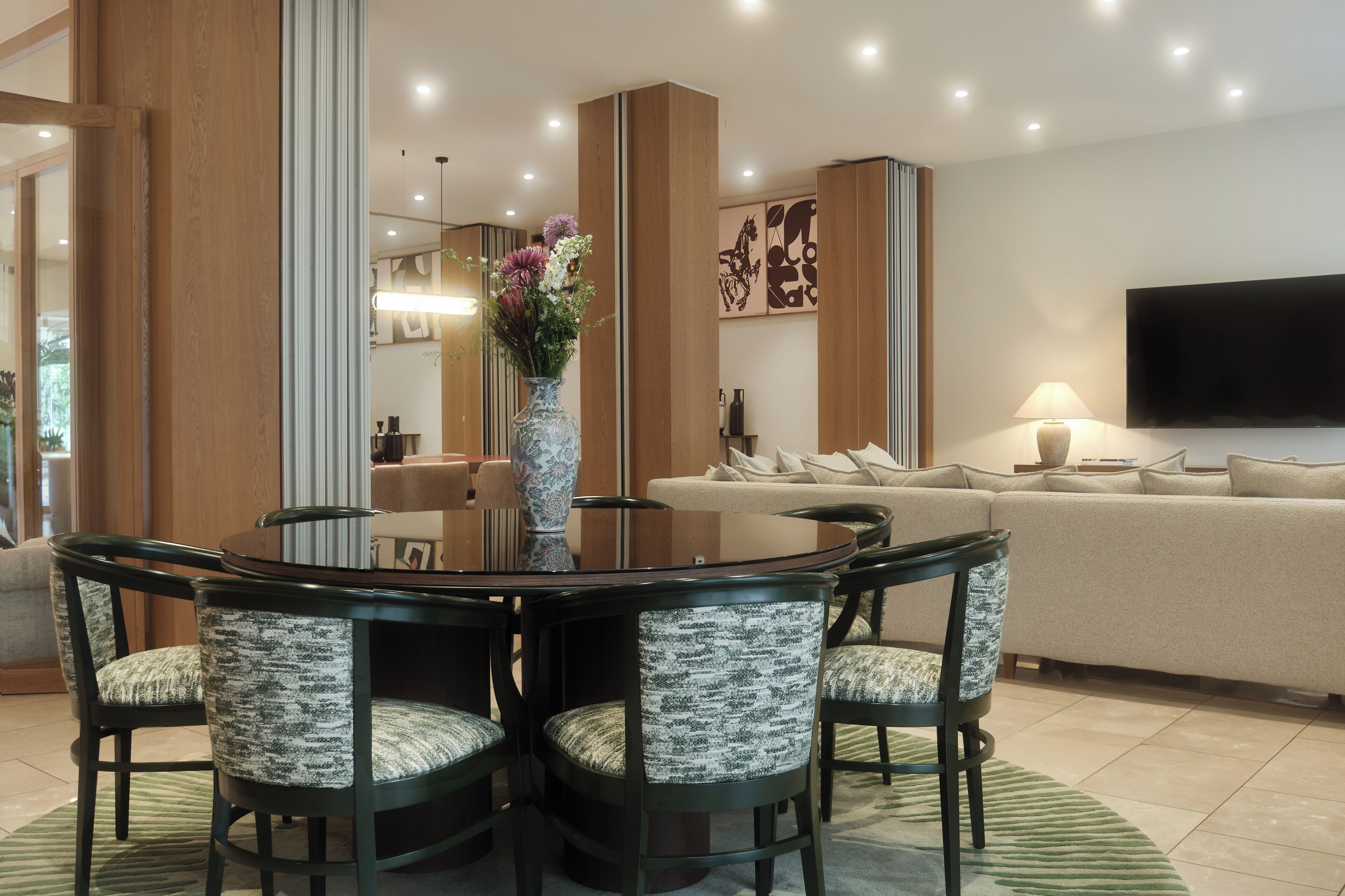 A modern living room with a round glass table, four upholstered chairs, a sofa, a flat-screen TV, and a vase with flowers.