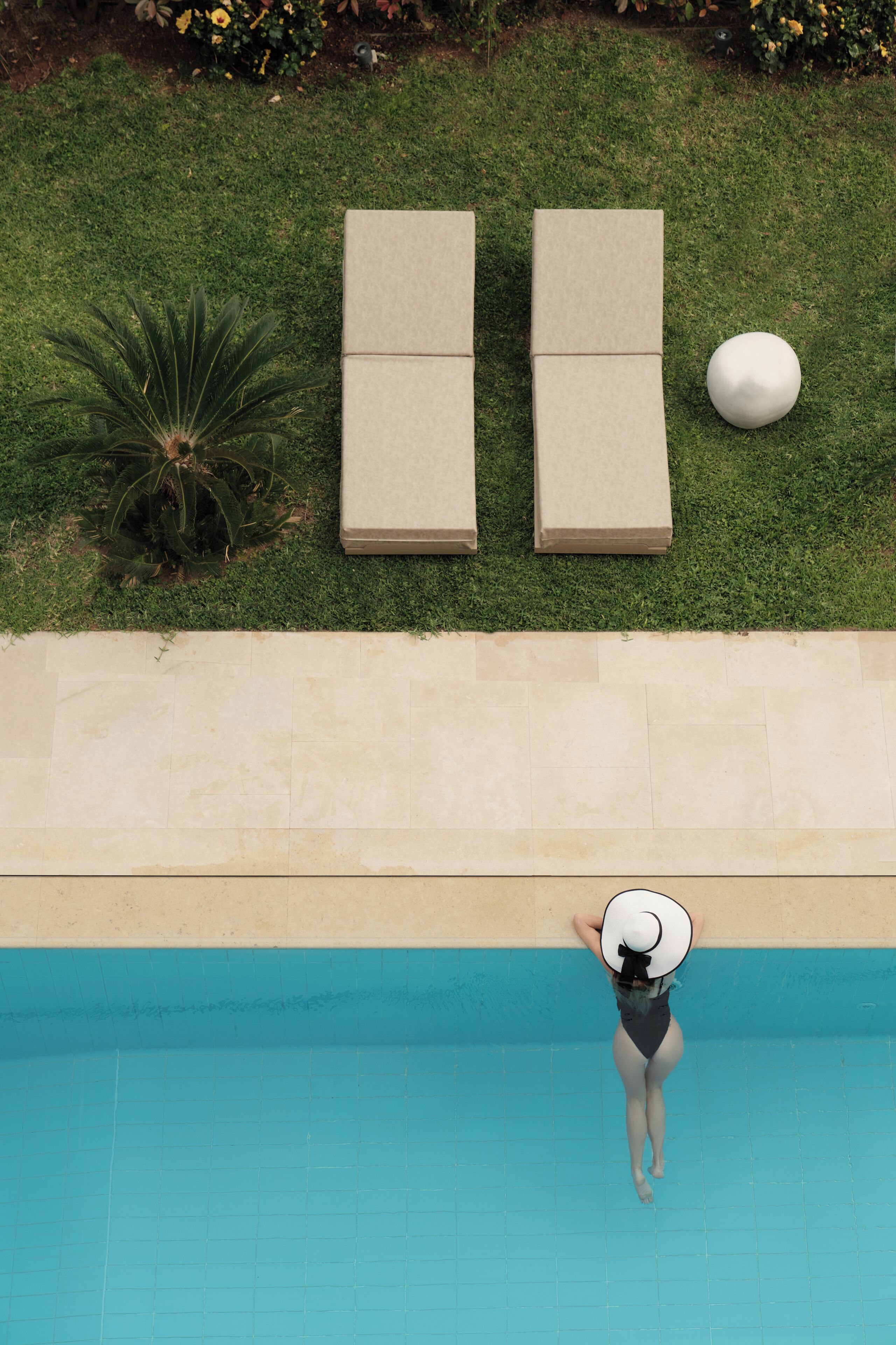 A person in a swimsuit standing at the edge of a swimming pool with lounge chairs and a plant on the grass.