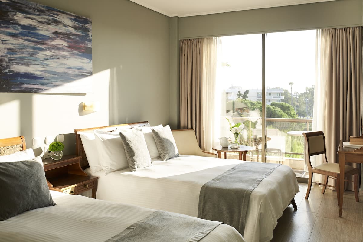 A hotel room with two beds, a large window with curtains, a desk with a chair, and a painting on the wall.
