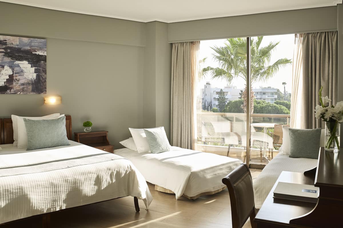 A hotel room with two beds, a desk, a chair, a lamp, and a large window with a view of palm trees and buildings.