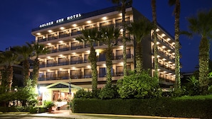 A multi-story hotel with balconies, palm trees, and a well-lit entrance. - Golden Sun (Glyfada)