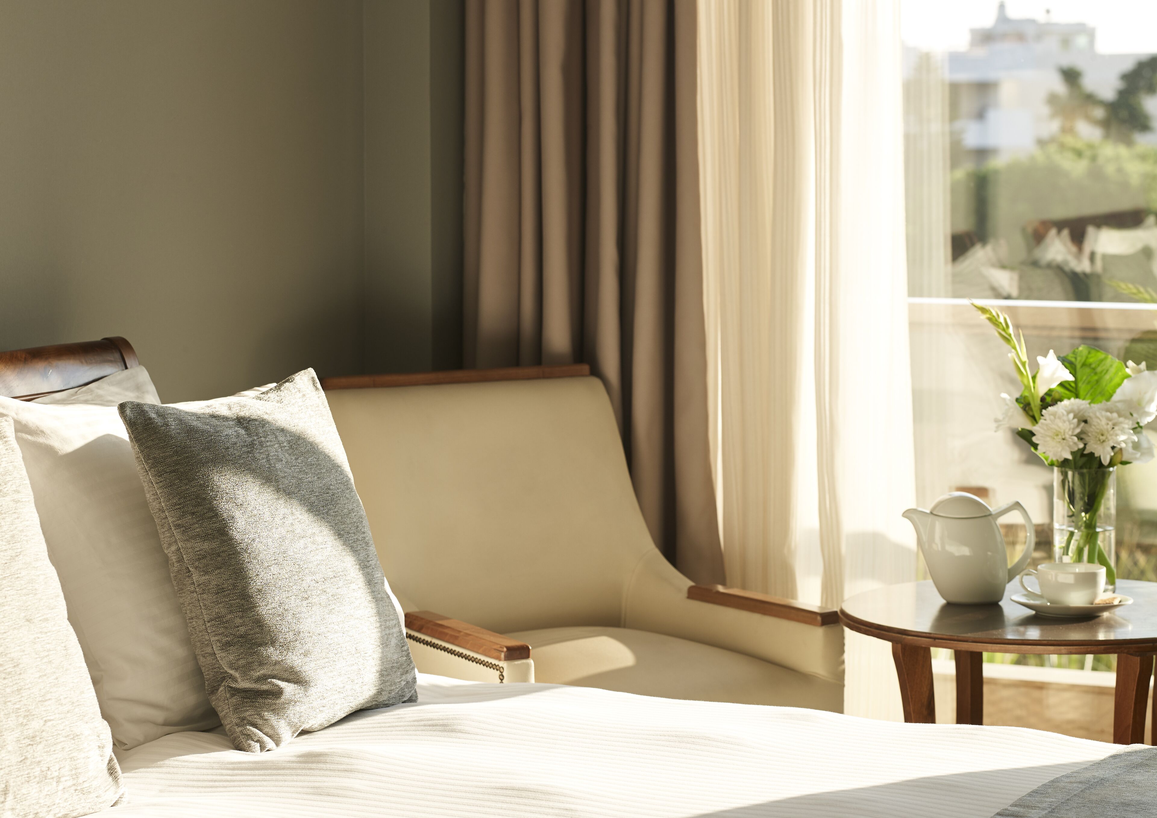 Habitación de hotel con cama, una silla, una mesa con tetera y flores, y vistas al exterior.