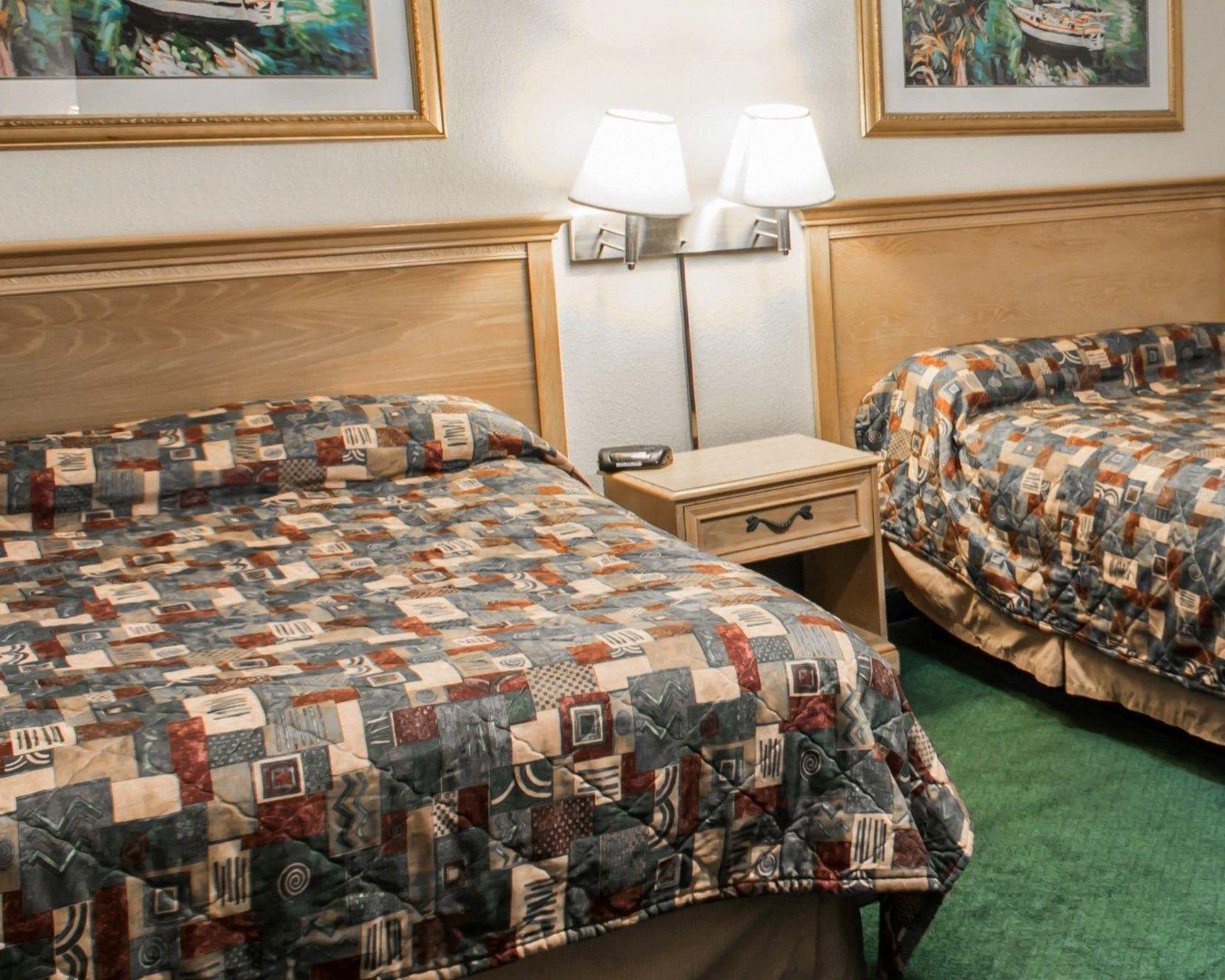 A hotel room with two beds, each with a quilted bedspread, a bedside table with a lamp, and a wall-mounted light.