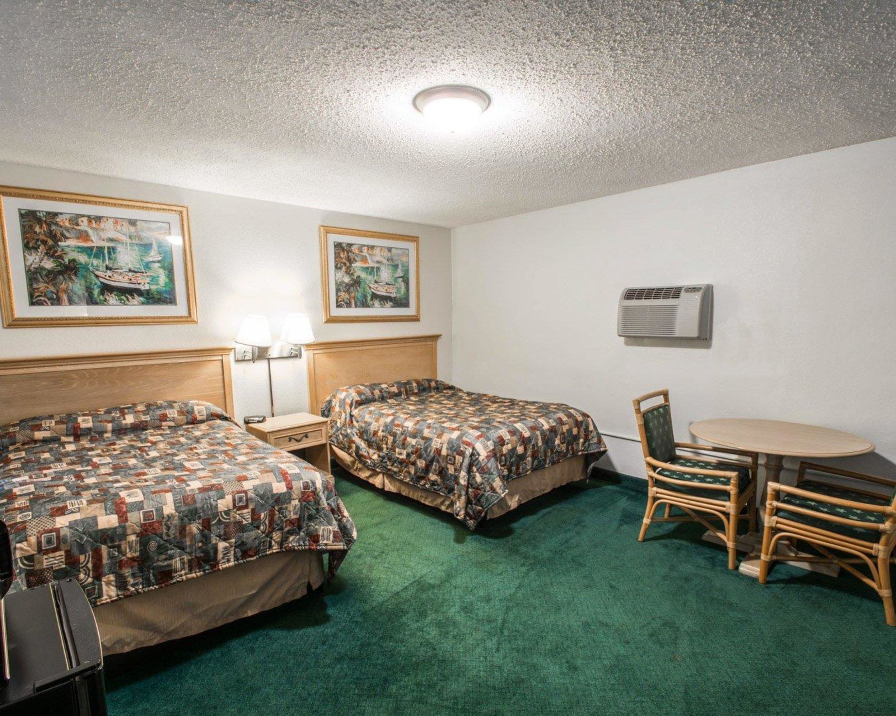 Quarto de hotel com duas camas, uma mesa pequena com cadeiras, um abajur e duas pinturas na parede.