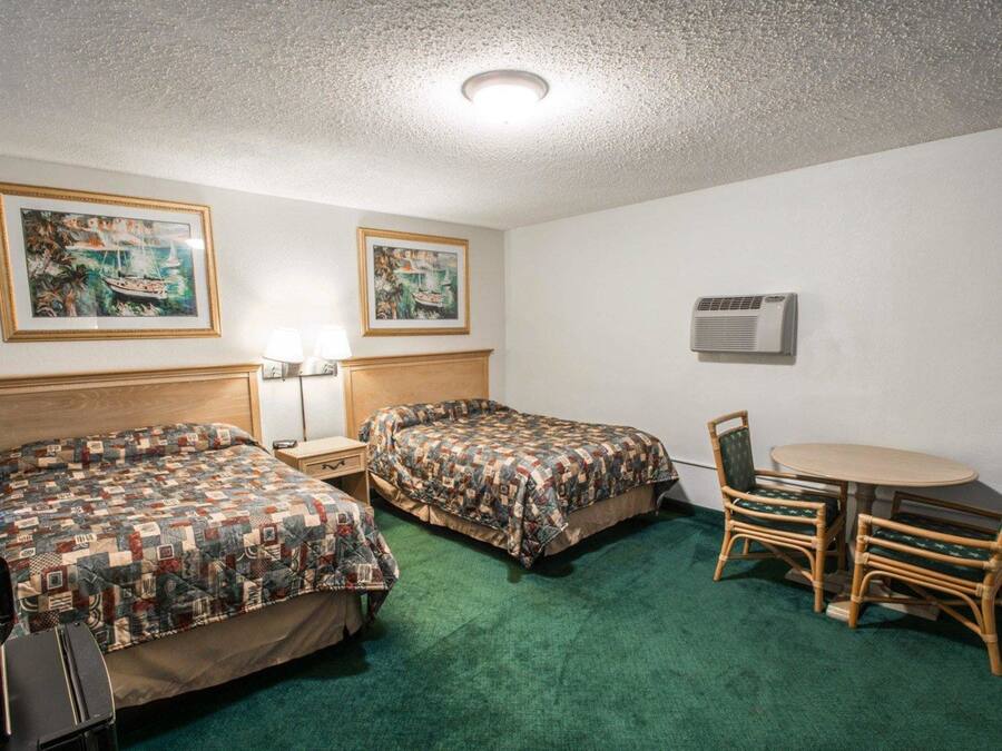 A hotel room with two beds, a small table with chairs, a lamp, and two paintings on the wall.