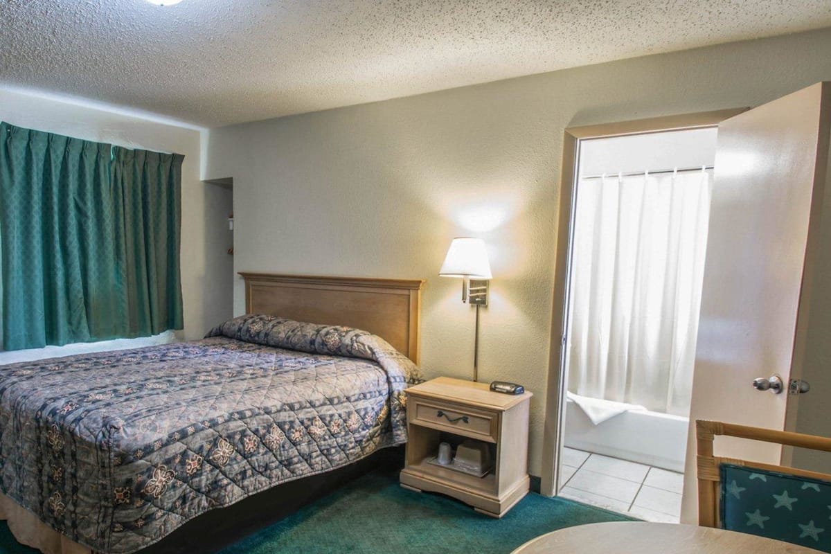 A hotel room with a bed, bedside table, lamp, and a door.