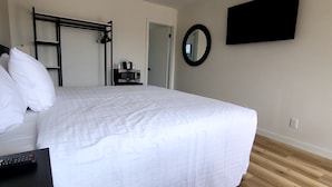 A neatly made bed with white bedding, a black bedside table with a remote control, a microwave on a small shelf, a black metal wardrobe, a round mirror on the wall, and a flat-screen TV mounted above the bed. - Richland Inn and Suites (Richland)