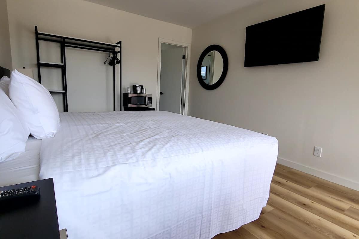 A neatly made bed with white bedding, a black bedside table with a remote control, a microwave on a small shelf, a black metal wardrobe, a round mirror on the wall, and a flat-screen TV mounted above the bed.