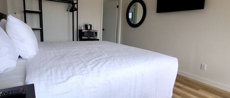 Una cama bien hecha con ropa de cama blanca, una mesita de noche negra con un control remoto, un microondas en un estante pequeño, un armario metálico negro, un espejo redondo en la pared y un televisor de pantalla plana montado sobre la cama.
