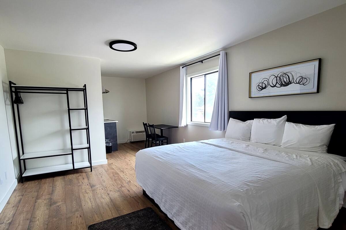 A bedroom with a large bed, a desk, and a chair. There is a window with curtains, a radiator, and a black mat on the floor.