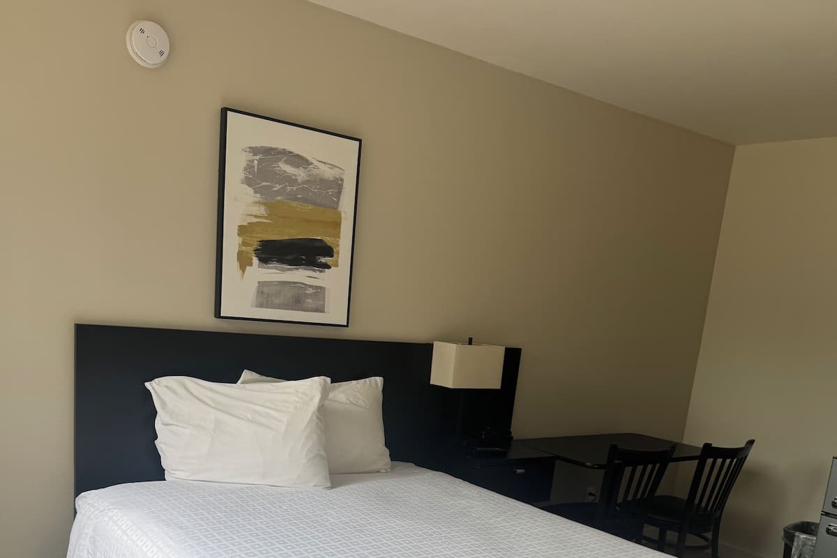 A neatly made bed with a white bedspread, a headboard, a bedside table with a lamp, and a framed abstract painting on the wall.