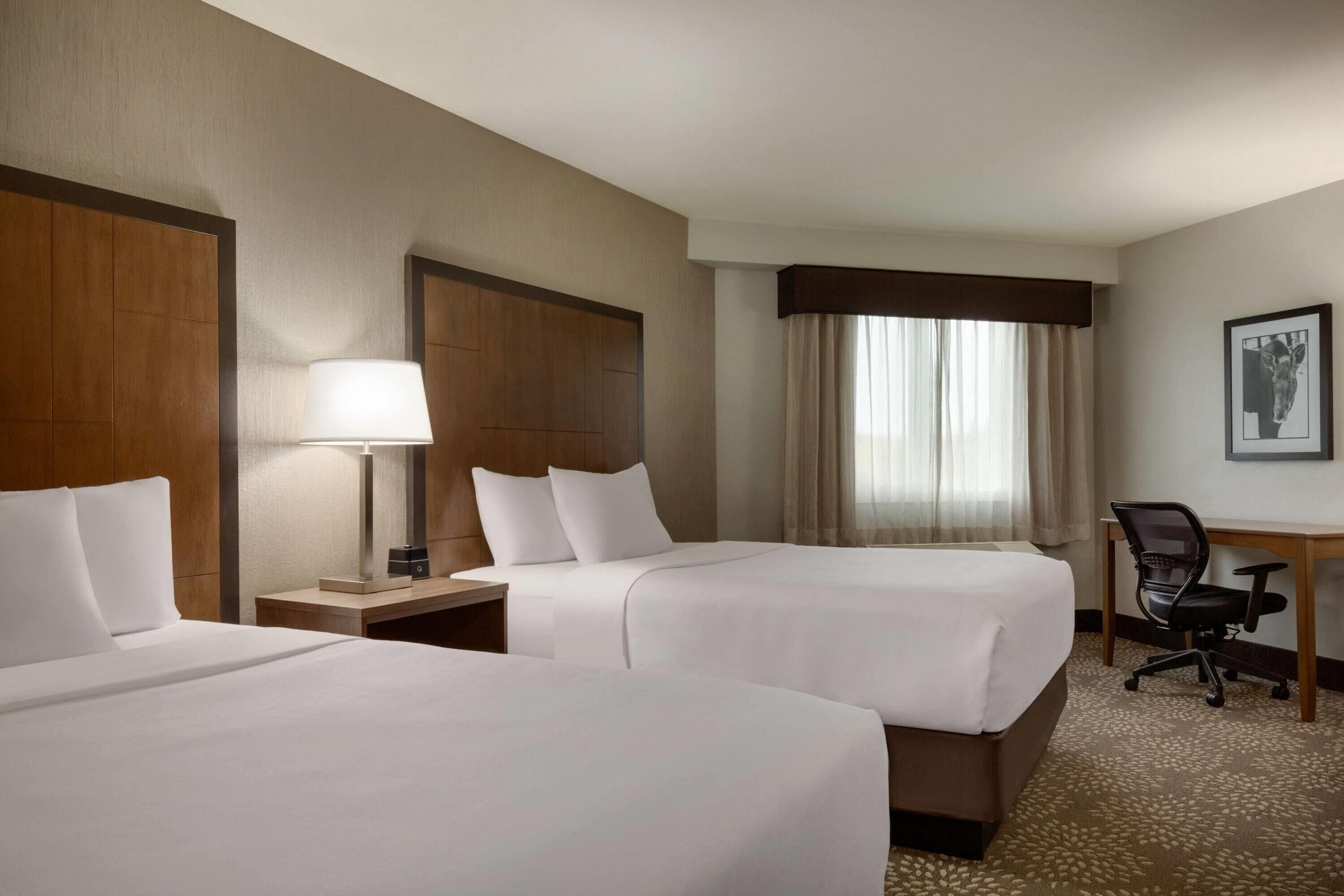 Quarto de hotel com duas camas, uma escrivaninha, uma cadeira, um abajur e uma janela com cortinas.