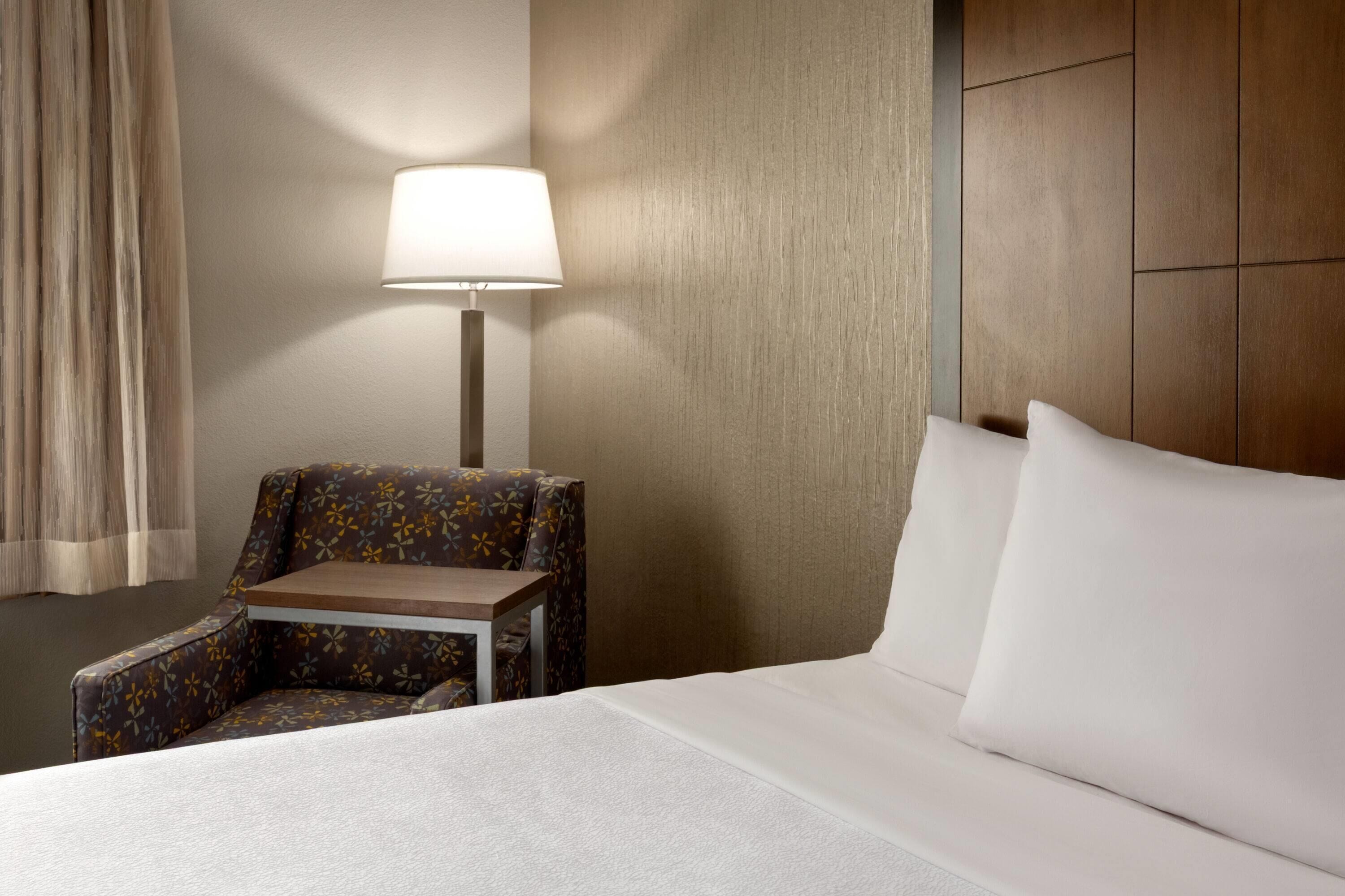 A hotel room with a bed, a chair, a small table, and a lamp.