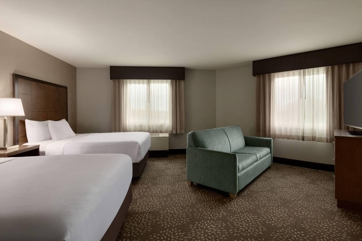 Une chambre d’hôtel avec deux lits, un fauteuil vert, une télévision et une fenêtre avec des rideaux.