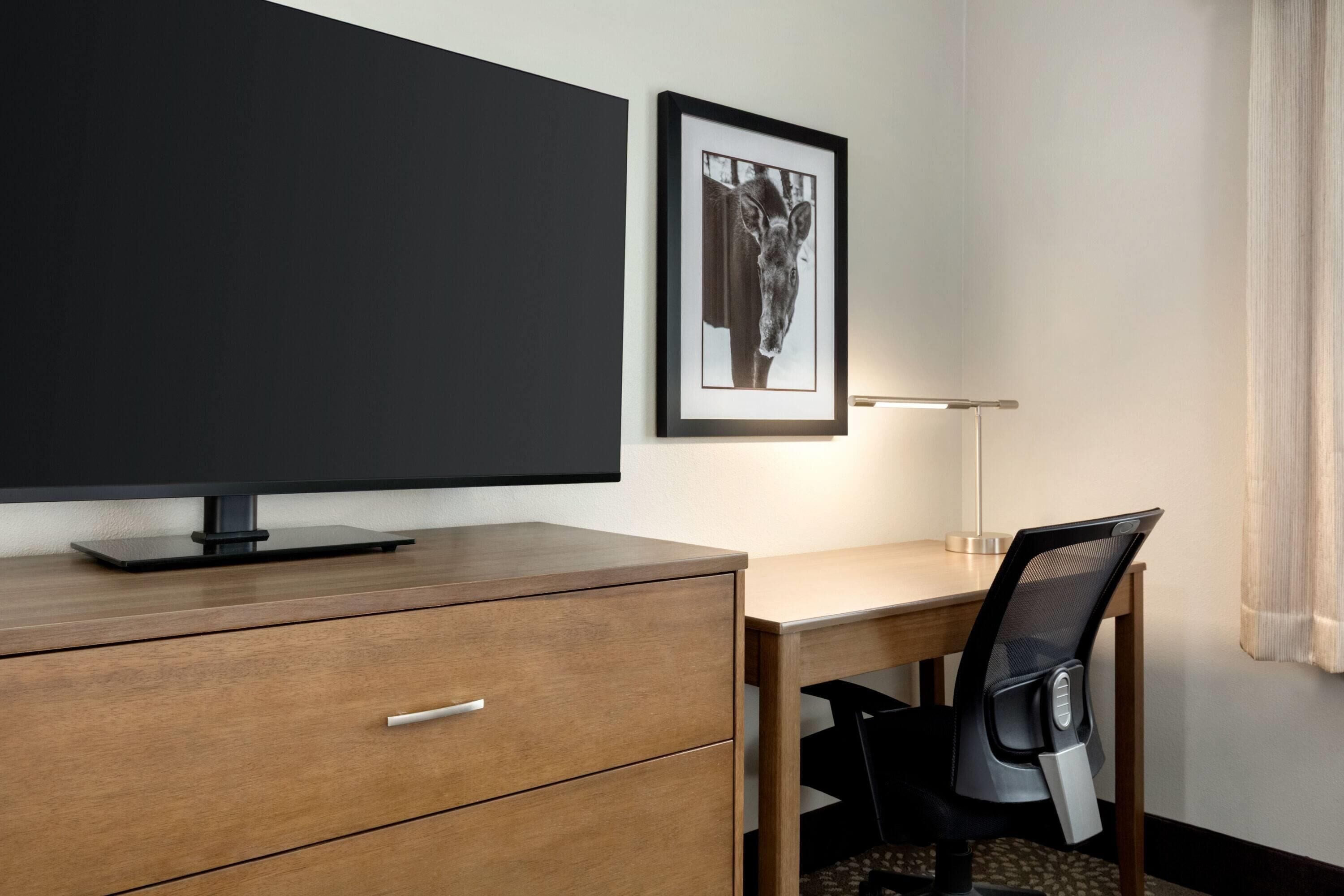 A hotel room with a wooden desk, a chair, a flat-screen TV on a stand, a framed picture on the wall, and a bedside lamp.