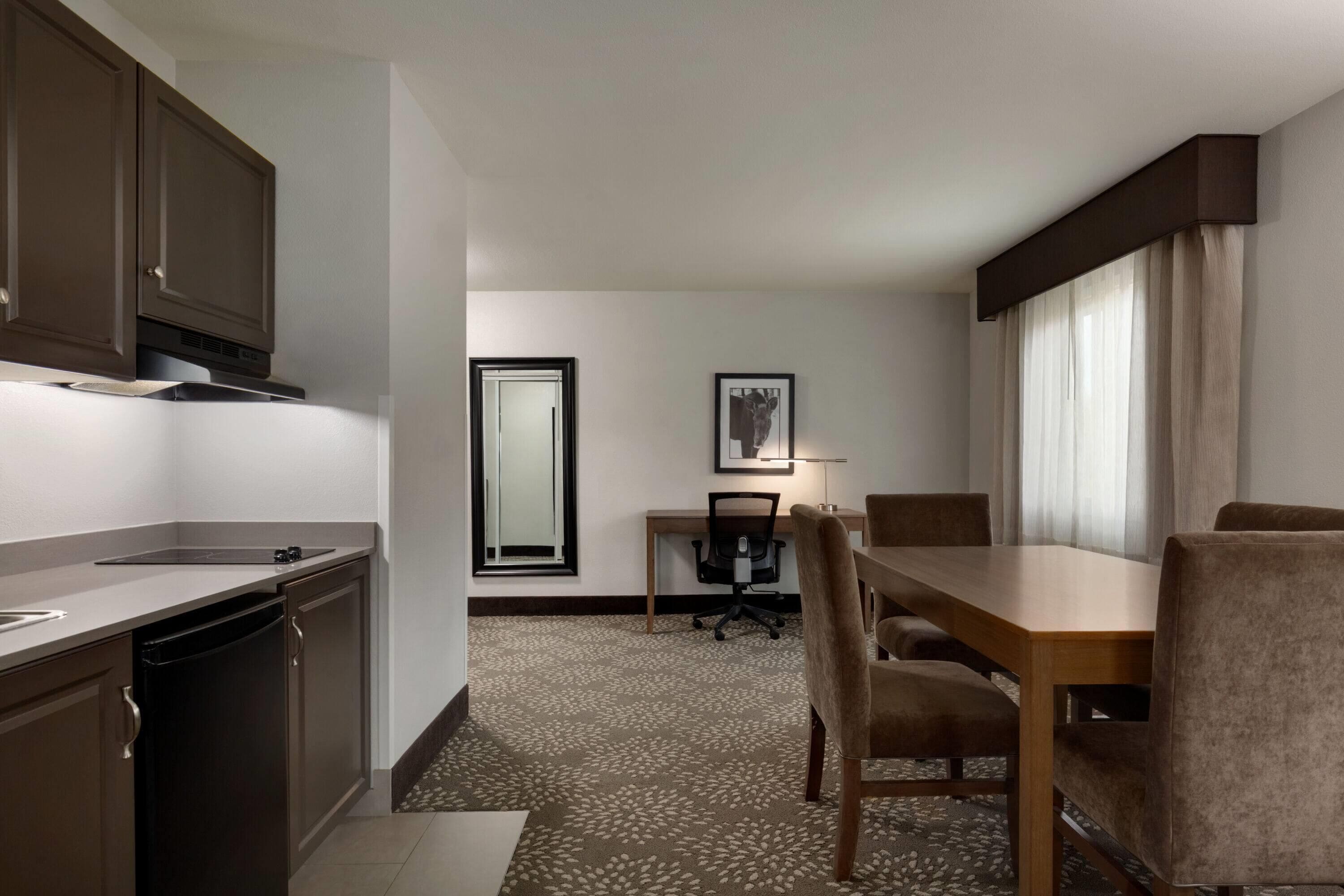 A compact hotel room with a kitchenette, a dining area with a table and chairs, a single office chair, and a large window with curtains.