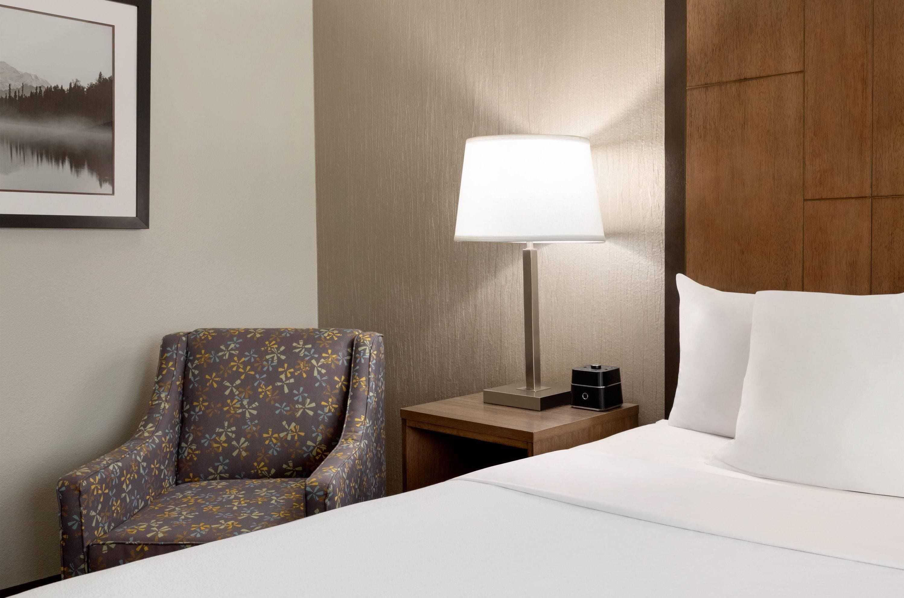 Una camera d'albergo con un letto, una sedia, un comodino con una lampada e un quadro appeso al muro.