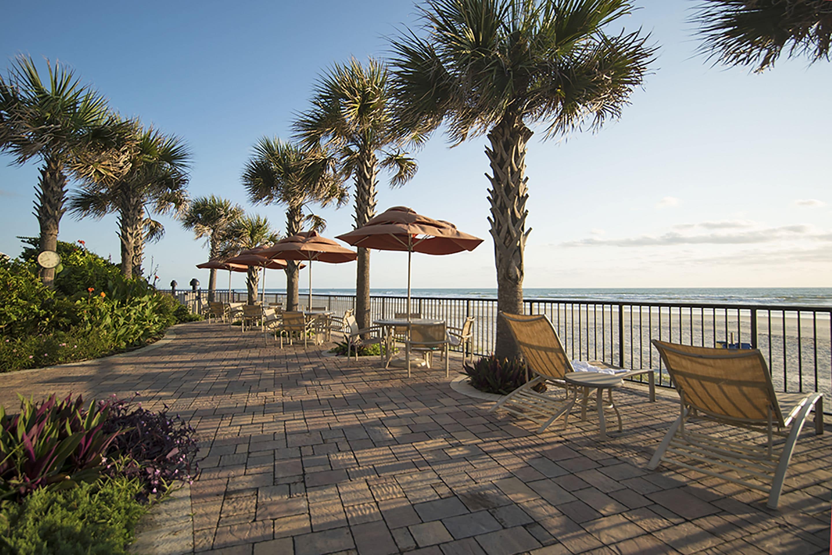 Ein gepflasterter Außenbereich mit Palmen, Liegestühlen und Sonnenschirmen mit Blick auf einen Strand.