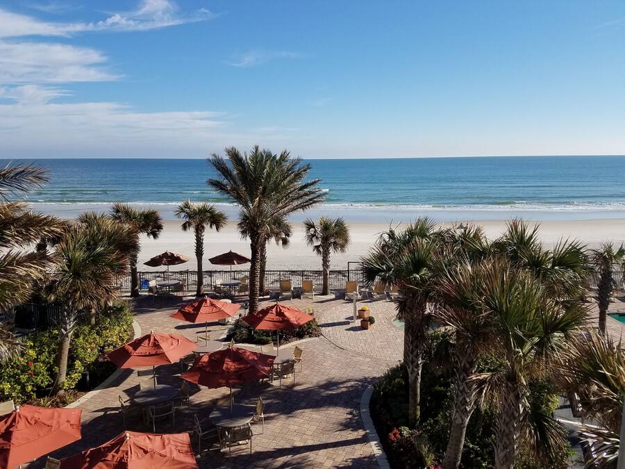 Un patio frente a la playa con mobiliario exterior y palmeras, con vista al océano.