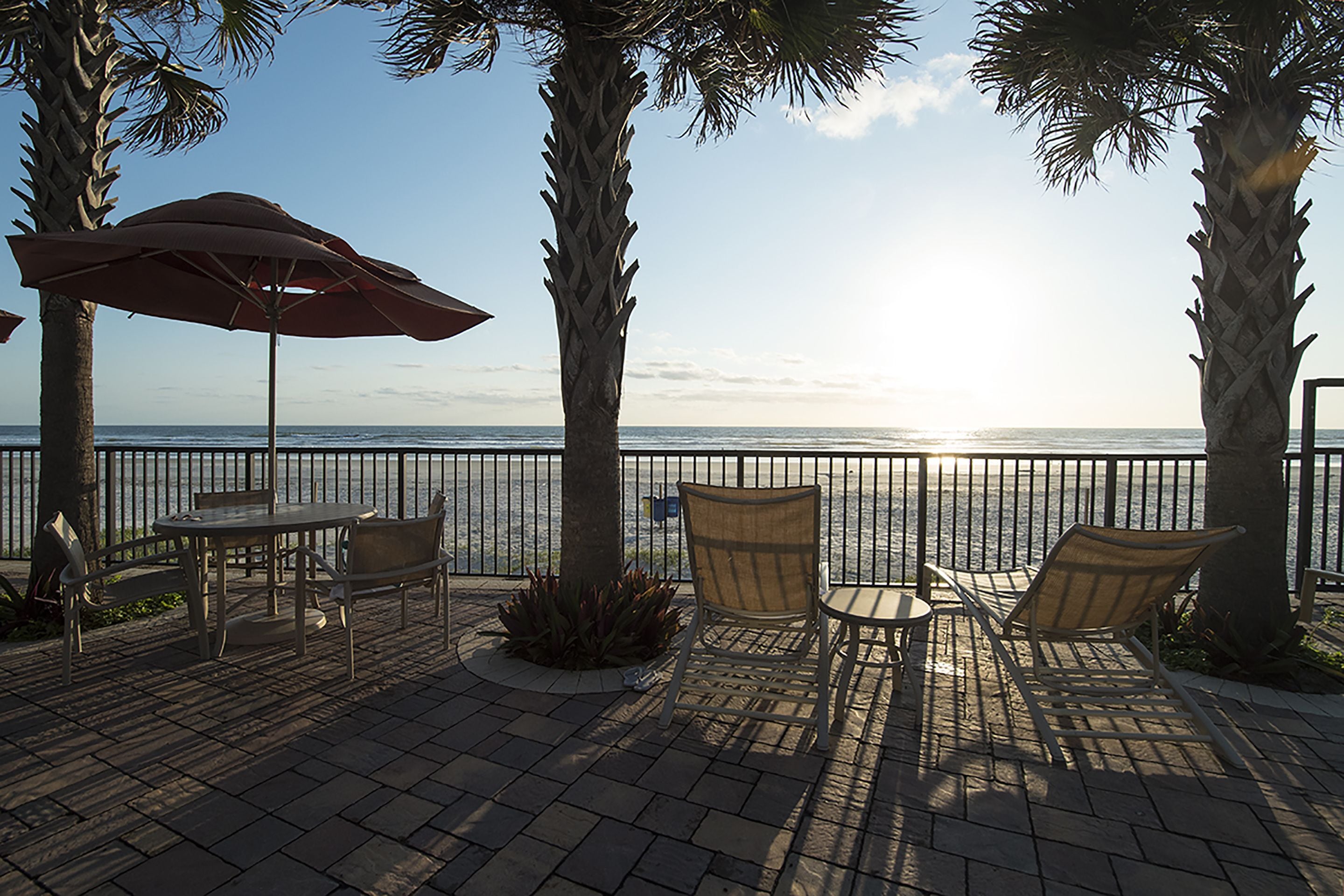 An outdoor patio with a table and chairs, a red umbrella, and a view of the ocean.