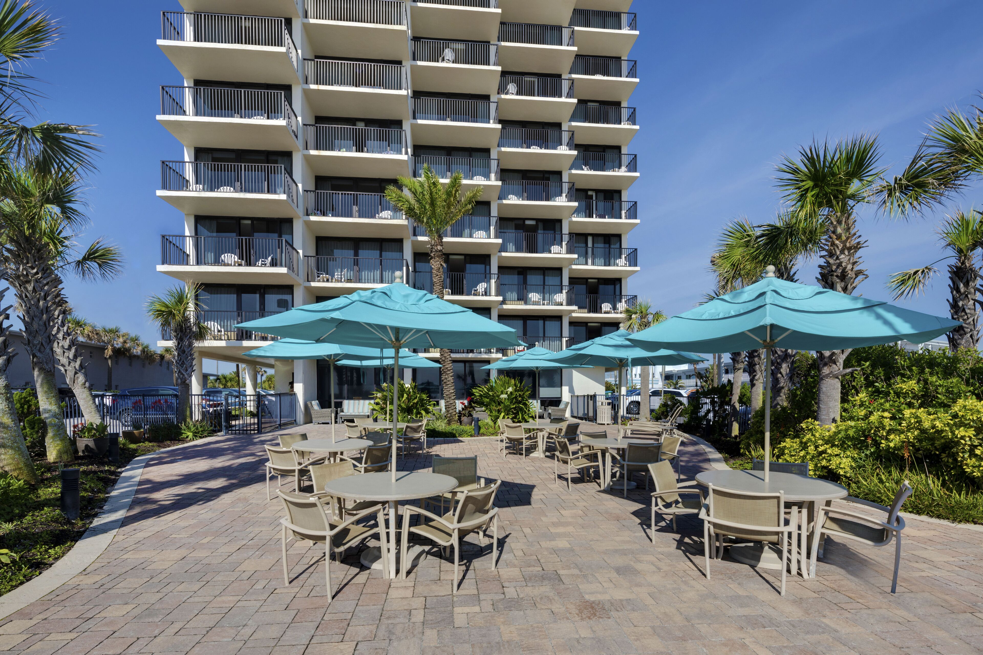 A hotel courtyard with outdoor seating and umbrellas, surrounded by palm trees and a multi-story building with balconies.