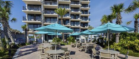 Un patio de hotel con mesas y sombrillas al aire libre, rodeado de palmeras y un edificio de varios pisos con balcones.
