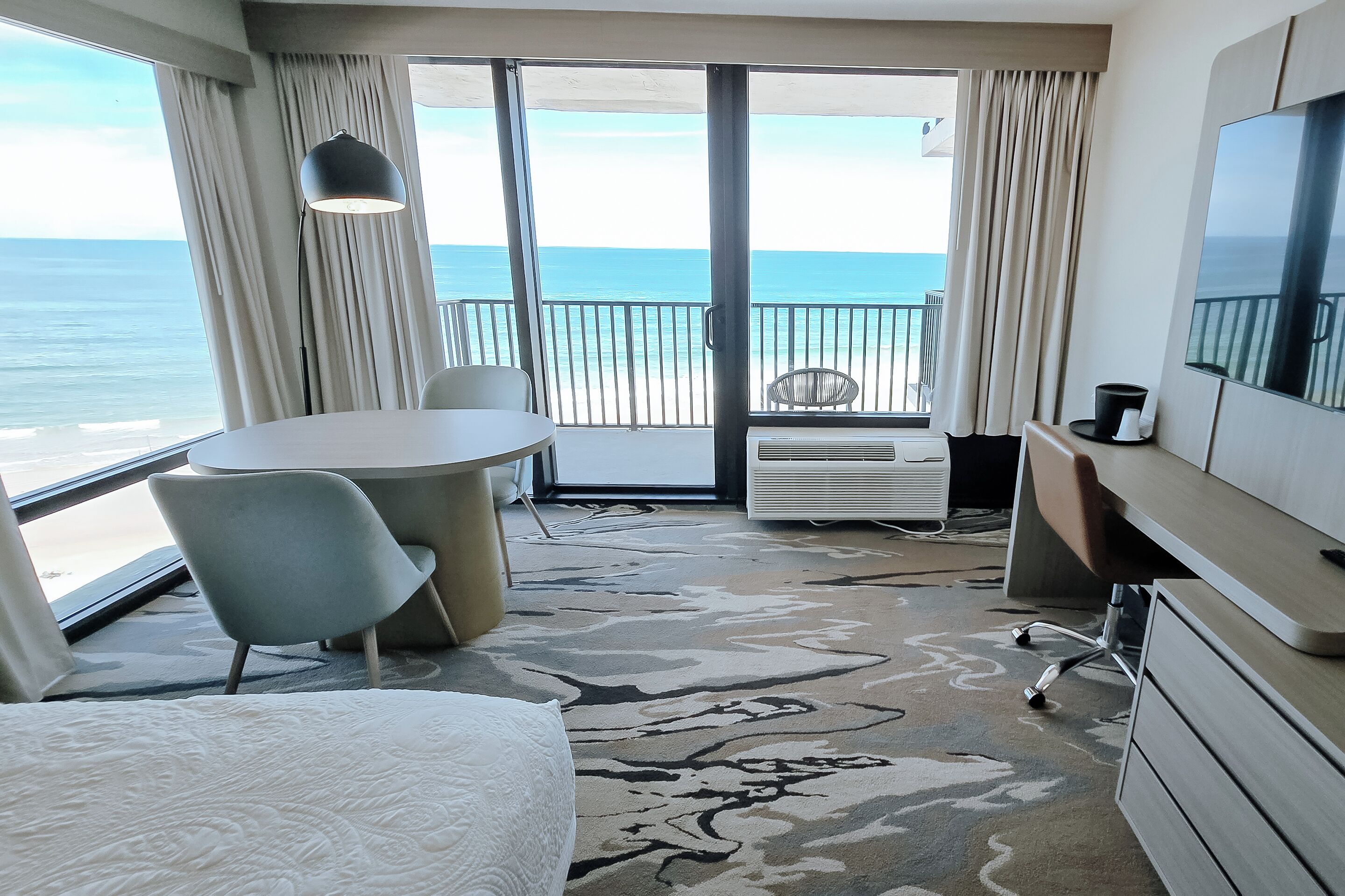 Habitación de hotel con cama, escritorio, silla, televisor y balcón con vista al océano.