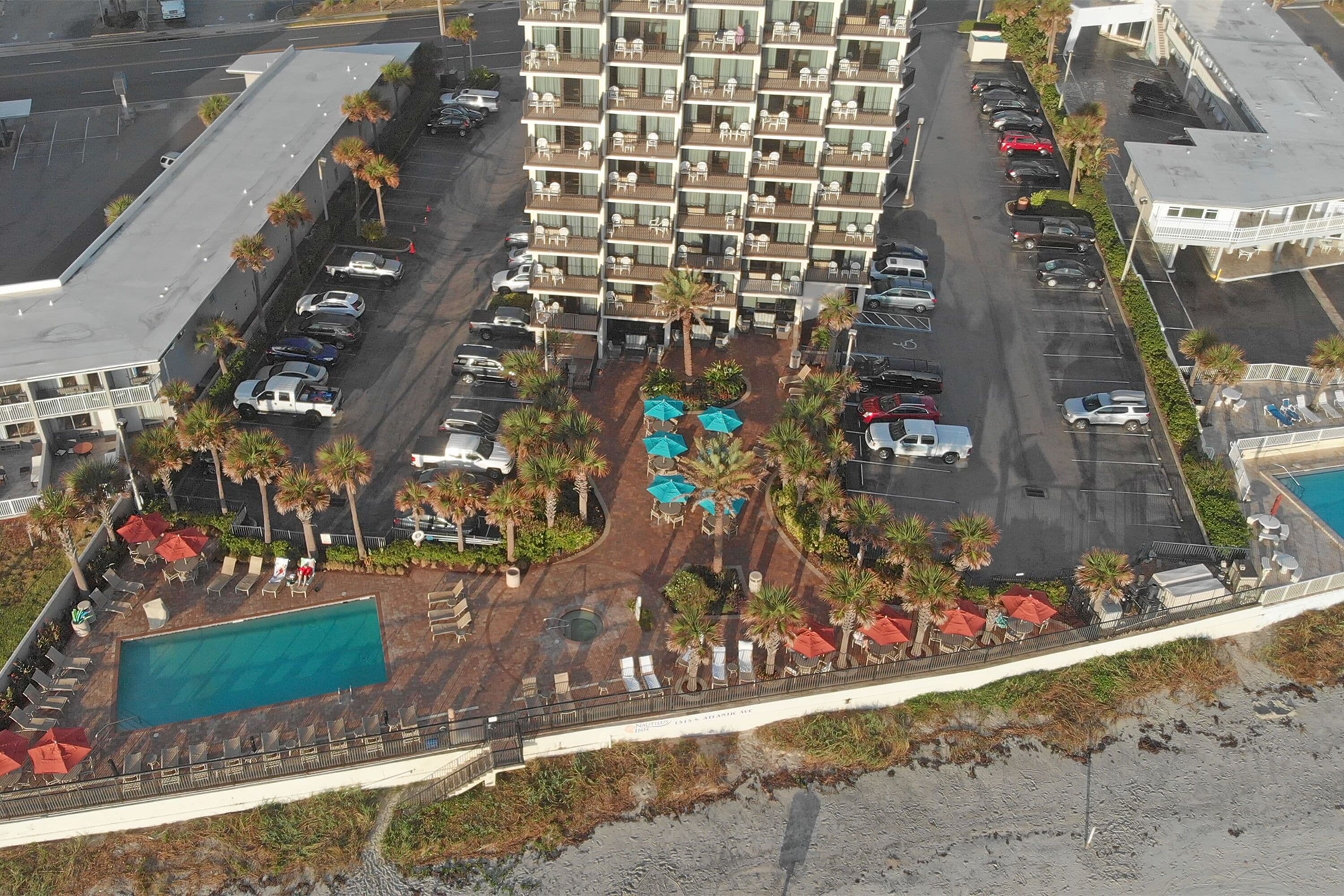 An aerial view of a hotel with multiple floors, a swimming pool, and a beach area with umbrellas.