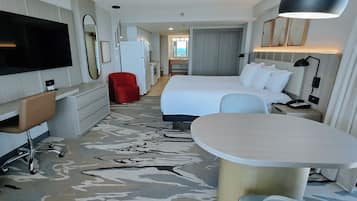 A modern hotel room with a large bed, a desk, a TV, a red chair, and a bathroom visible in the background.