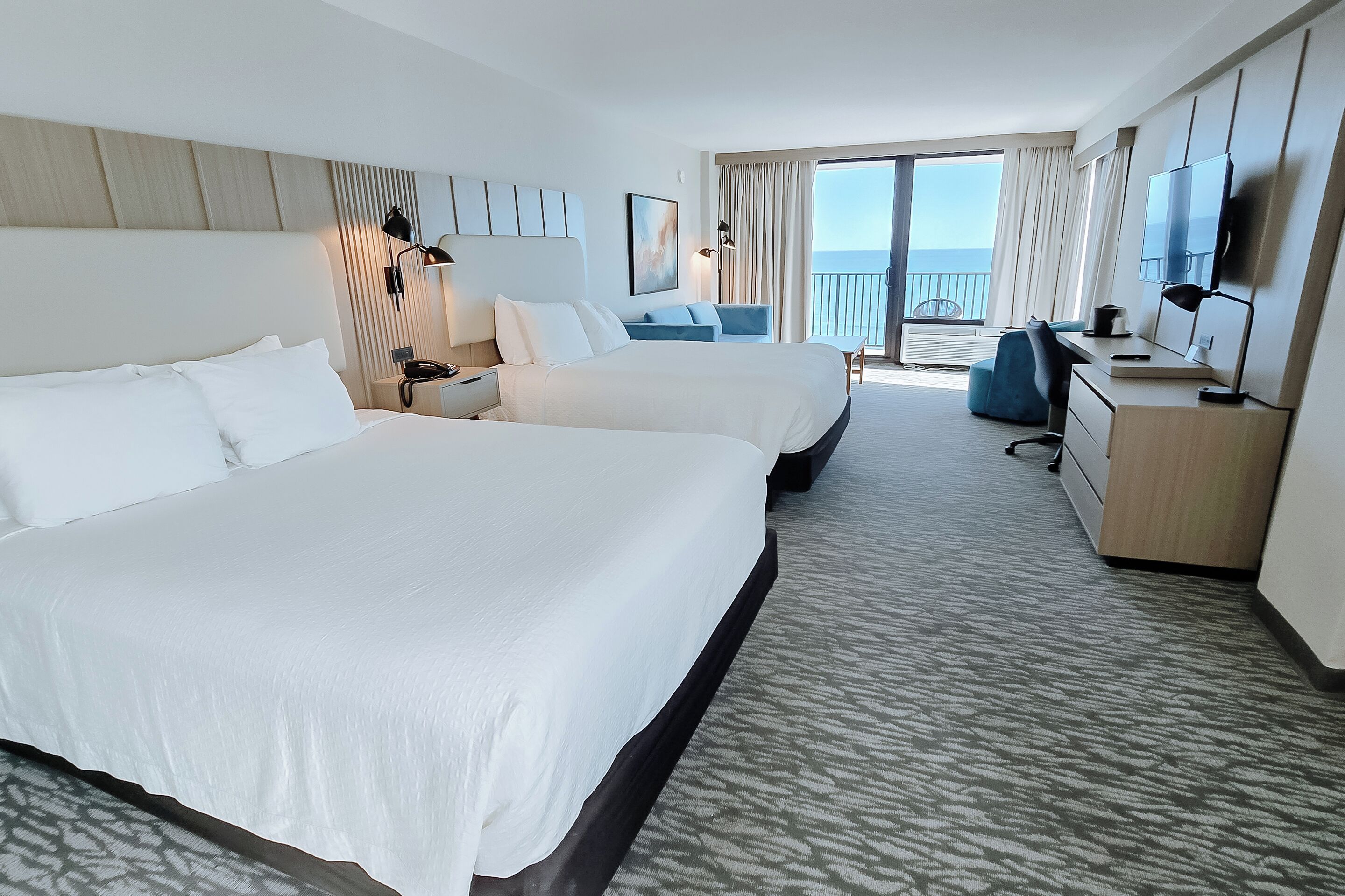 Une chambre d’hôtel avec deux lits, un bureau, une chaise et un balcon donnant sur la mer.