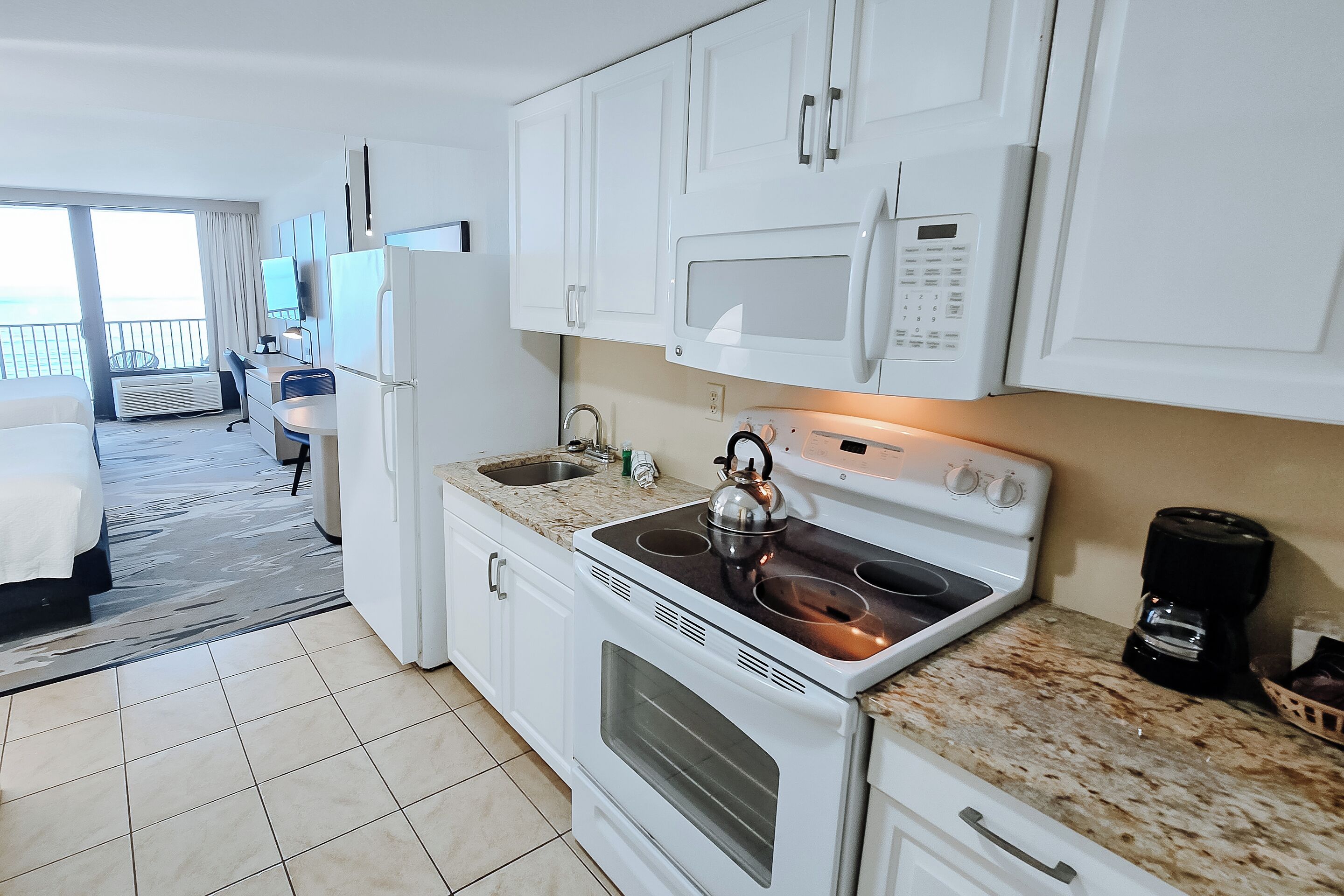 A compact kitchen with a stove, microwave, and sink. A kettle is on the stove, and a coffee maker is on the countertop.