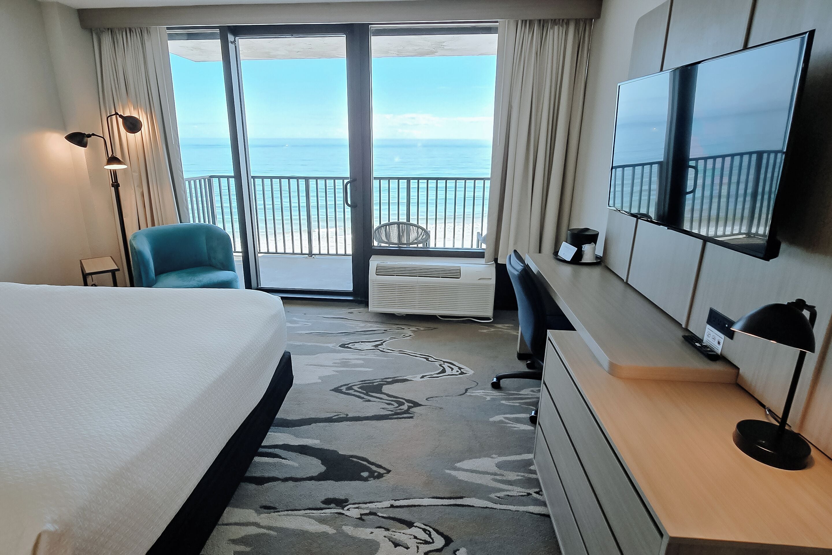 A hotel room with a bed, a desk, a chair, a TV, and a view of the ocean through a sliding door.