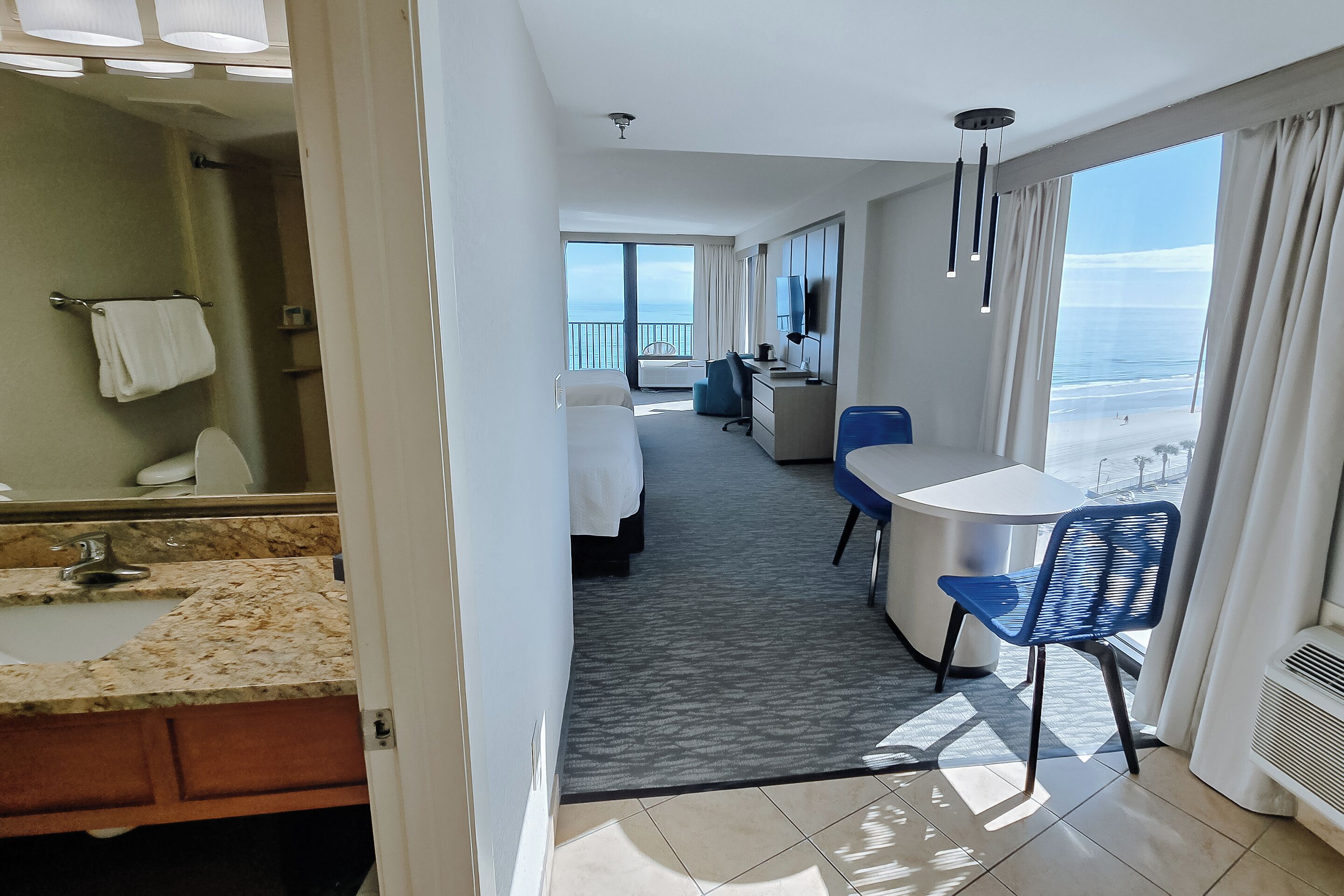 A hotel room with a bed, a desk, a chair, a bathroom with a sink and mirror, and a large window with a view of the ocean.