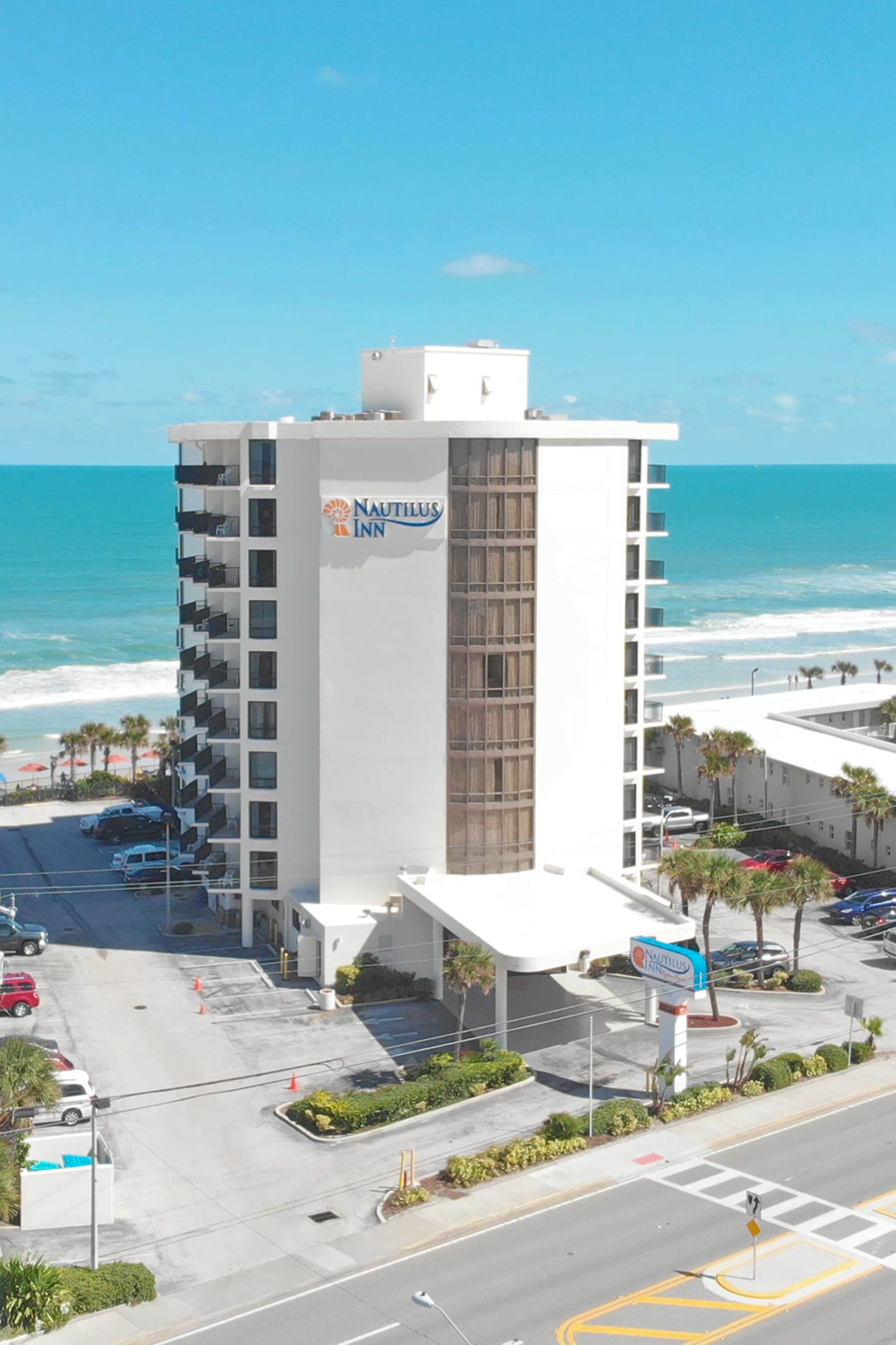 A multi-story hotel with a prominent sign for Nautilus Inn, located near a beach with visible parking and a road in the foreground.