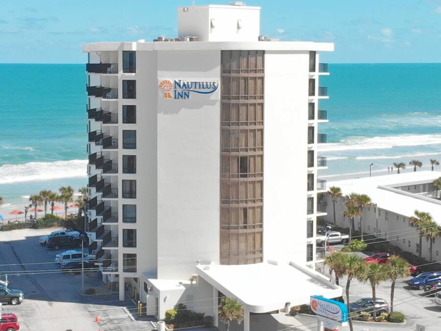 A multi-story hotel with a prominent sign for Nautilus Inn, located near a beach with visible parking and a road in the foreground.
