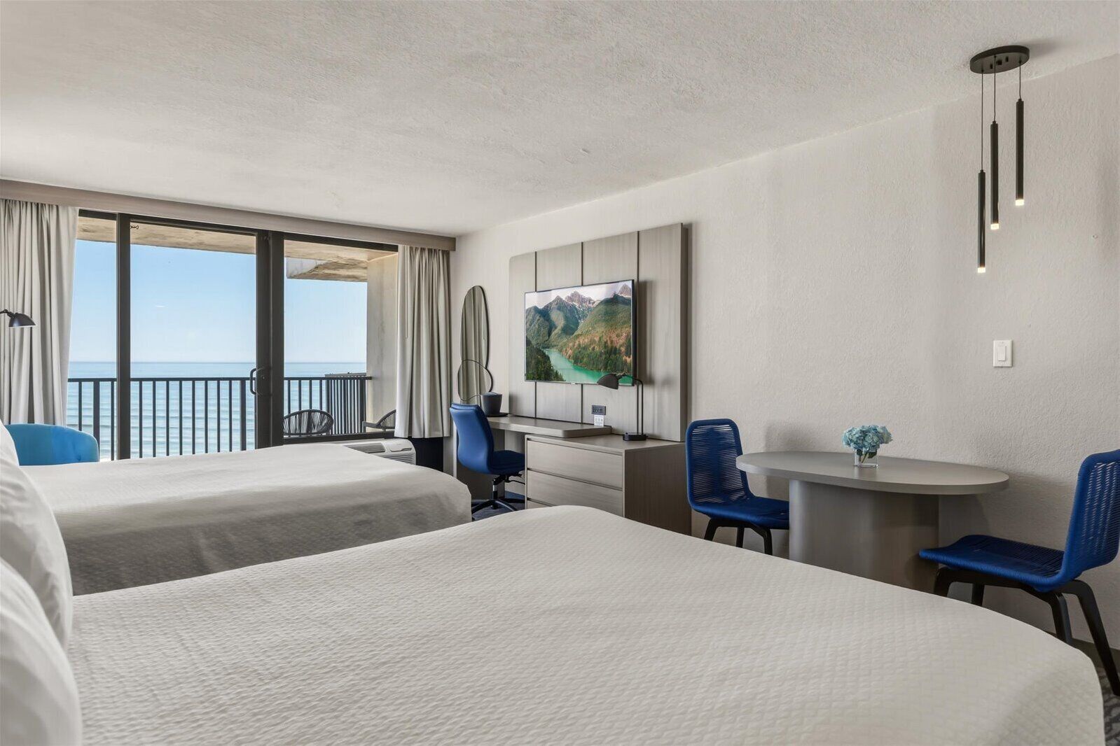 A hotel room with two beds, a desk with a chair, a small table with a chair, a TV, and a balcony with a view of the sea.