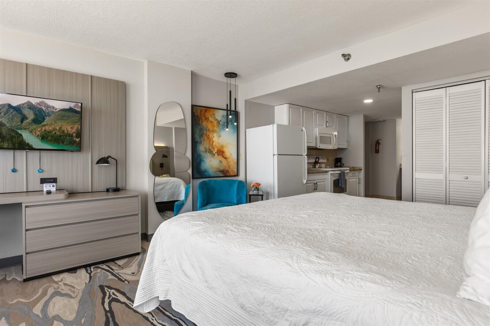 A hotel room with a bed, a desk, a TV, a painting, a kitchen area, and a wardrobe.