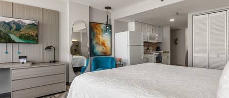 A hotel room with a bed, a desk, a TV, a painting, a kitchen area, and a wardrobe.