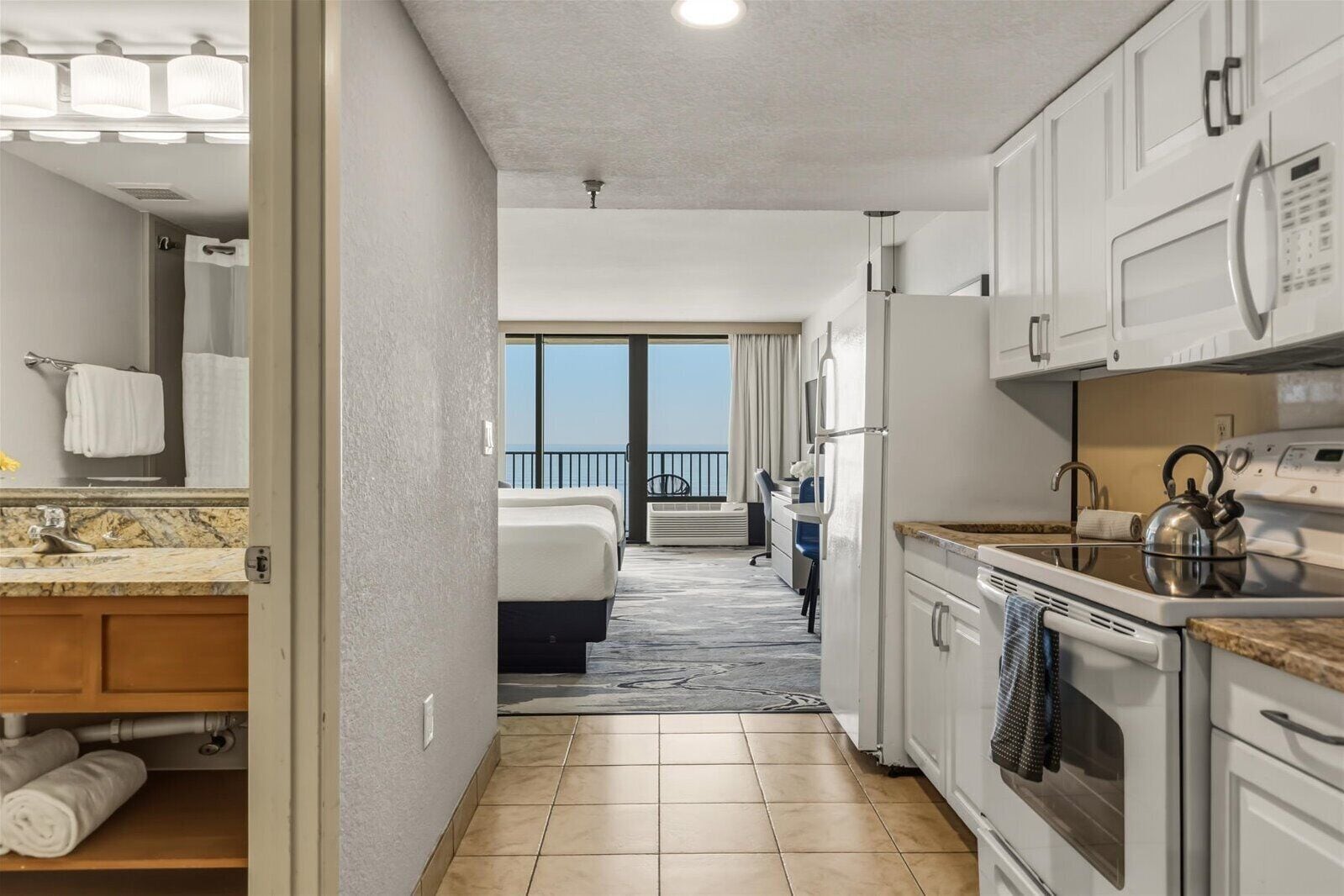 A hotel room with a bathroom, kitchenette, and a balcony with a view.