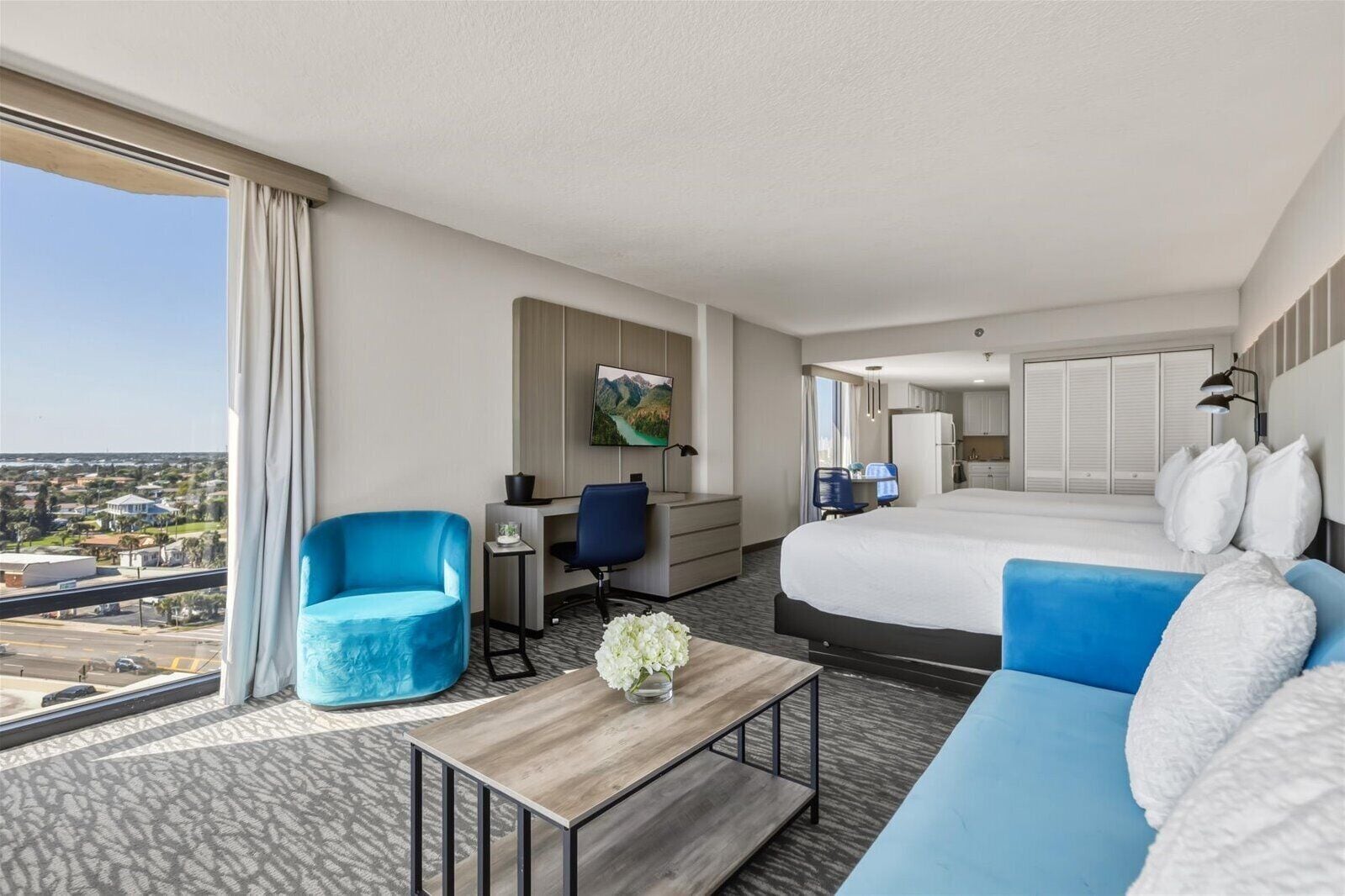 A hotel room with a large bed, a blue armchair, a desk with a chair, a small table with a vase, and a view of the city.