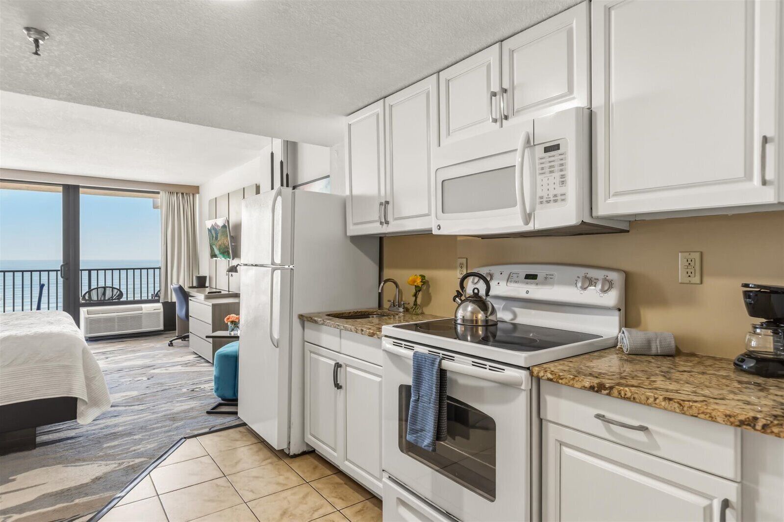 A modern kitchen with white cabinets, a microwave, stove, and a kettle. There's a balcony with a view.
