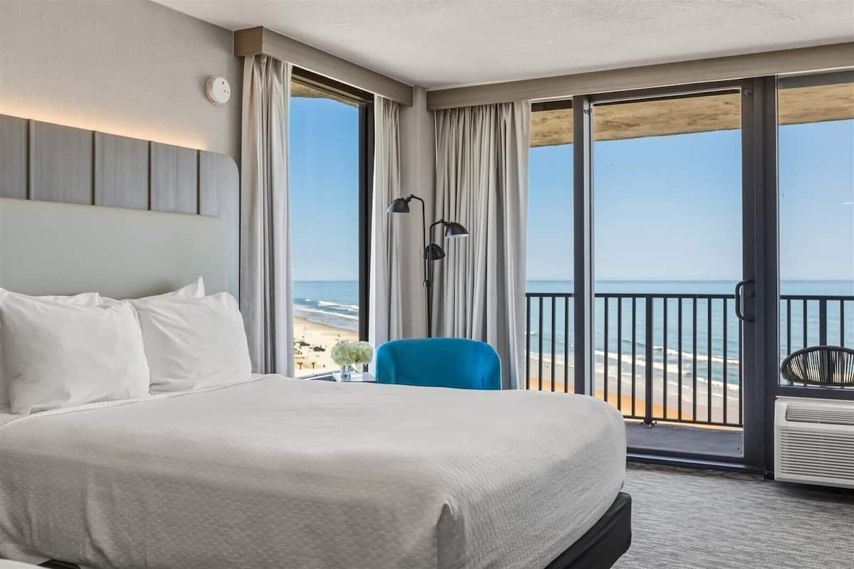 A hotel room with a large bed, a blue chair, a nightstand, a lamp, and a view of the beach.