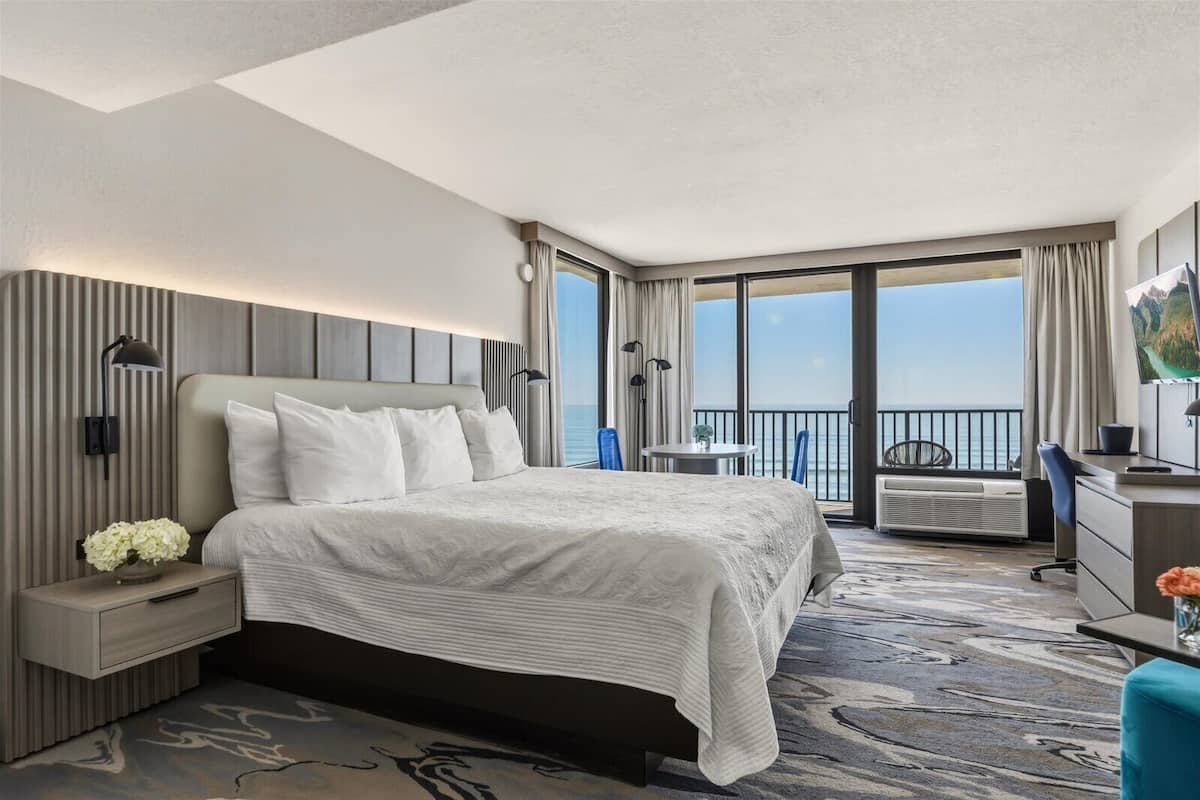A hotel room with a large bed, a desk, a chair, a small table, and a balcony with a view of the ocean.