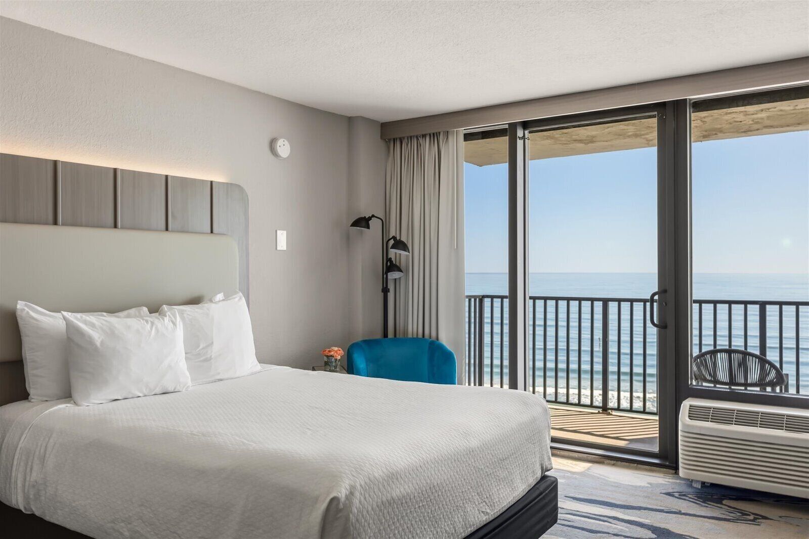 A hotel room with a large bed, a chair, a desk, a lamp, and a view of the ocean.