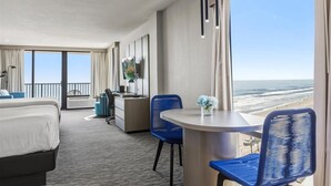 A hotel room with a bed, desk, chairs, a TV, and a large window with a beach view.