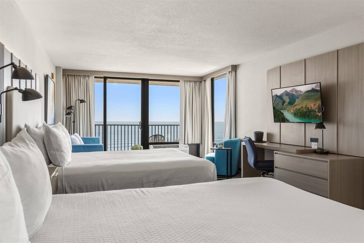 A hotel room with two beds, a desk, a chair, a TV, and a view of the sea.
