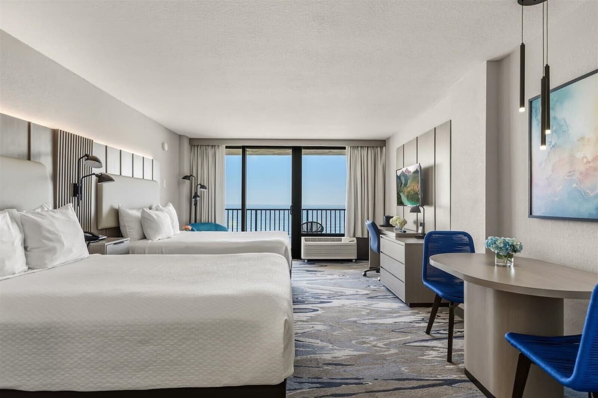 A hotel room with two beds, a desk, chairs, a large window with a view, and a painting on the wall.