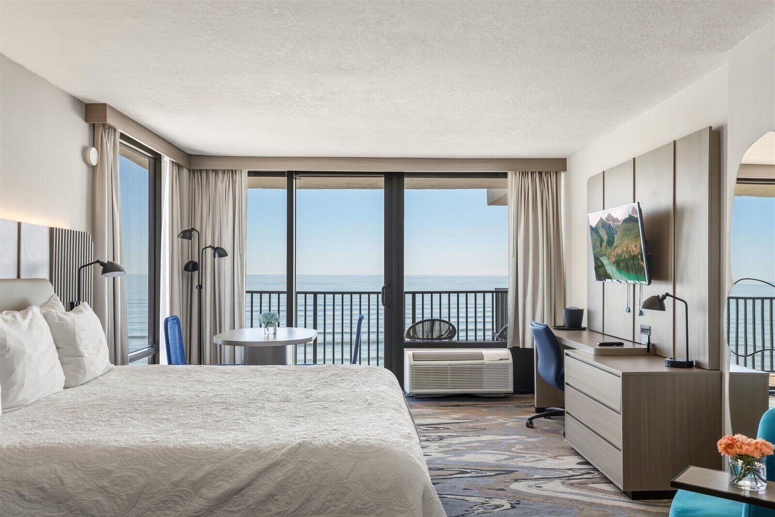 A hotel room with a large bed, a desk, a chair, a TV, and a view of the sea.
