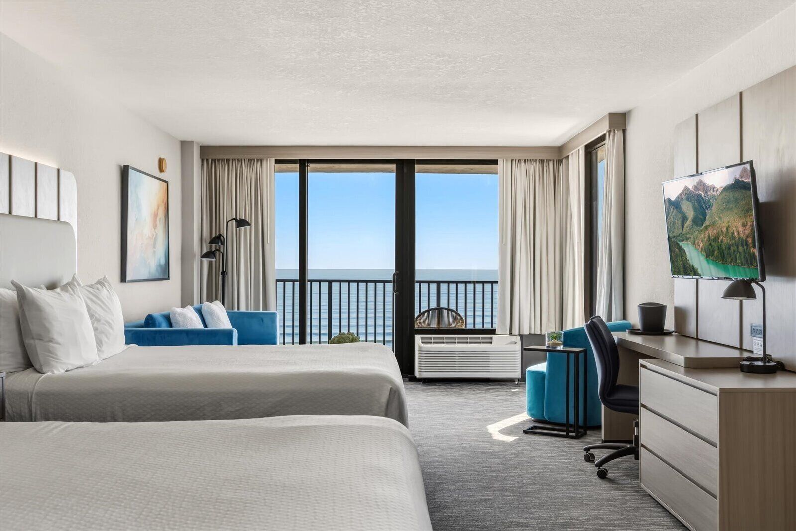 A hotel room with two beds, a desk, a chair, a TV, and a balcony with a view of the sea.