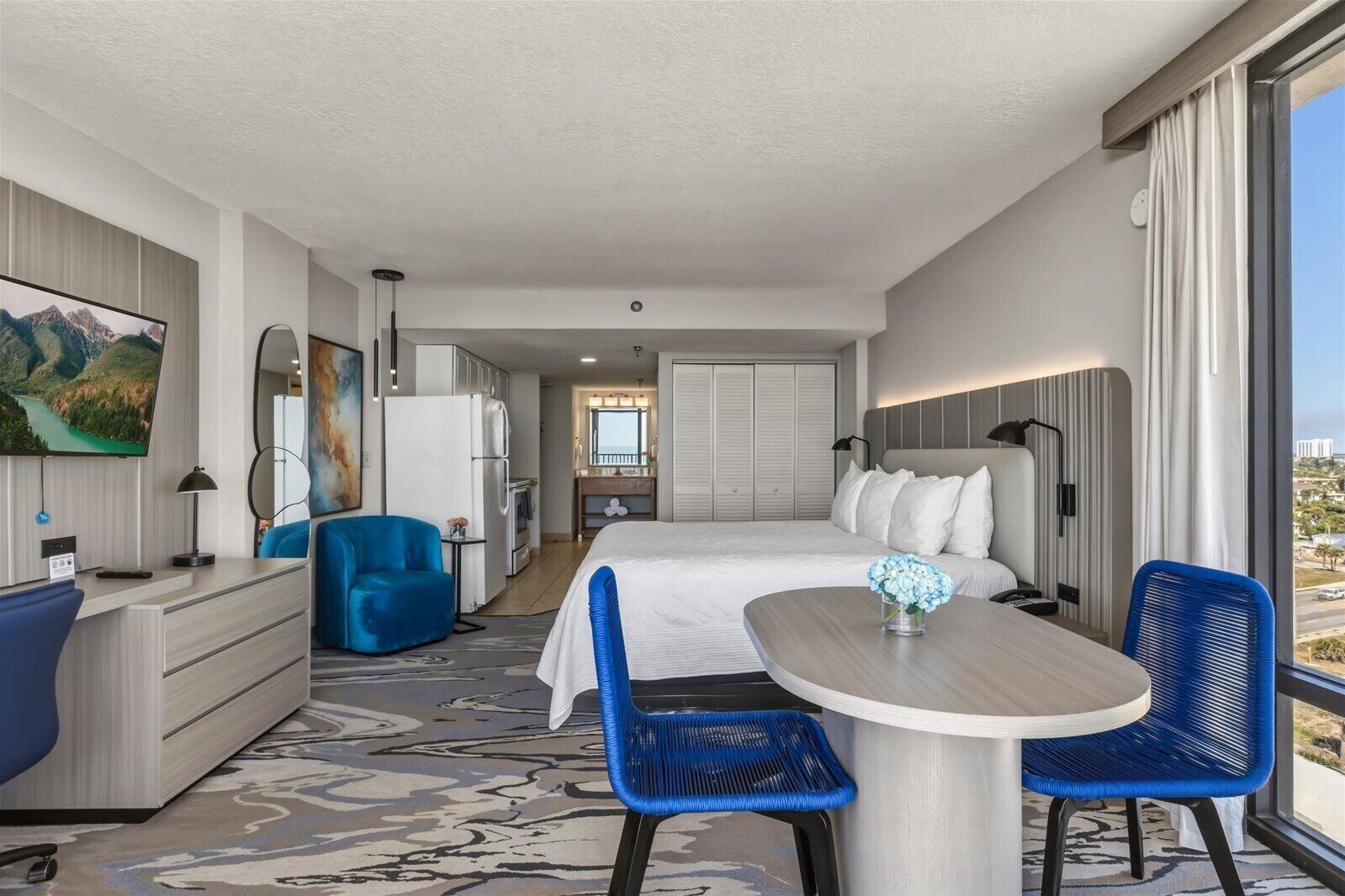 A hotel room with a bed, a desk, a chair, a table, a TV, and a view of the city.